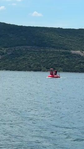 The video shows a small, red, flat-bottomed boat moving across a body of water. There are at least two people on the boat. In the background is a hilly, green landscape. The sky is blue with some white clouds. The water is choppy.