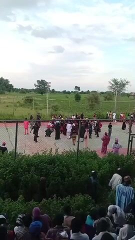 The video shows a group of people gathered outdoors. In the foreground, a crowd of spectators is seated, many wearing traditional headwear. Behind them, a fenced-in court, possibly a basketball or tennis court, is visible. Several individuals, dressed in various colorful attire, are interacting and moving around on the court. In the background, there is a vast expanse of green fields and scattered trees under a cloudy sky. The overall atmosphere appears to be one of a community gathering or event.