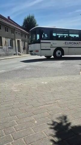 A white bus with "EUROMATPOL" printed on its side drives along a paved road. The bus is white with black tinted windows. A residential building with a tiled roof is visible on the left side of the road, behind a sidewalk made of brick pavers. The sky is blue with scattered clouds, and a tree is visible behind the residential building. Further down the road, a white van and another vehicle are traveling in the same direction as the bus. The van has text and logos on its side. The scene appears to be a sunny day in a town or city.