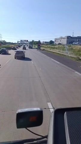 The video shows a view from a vehicle driving on a highway in Irxleben, Germany. The highway has multiple lanes and is lined with grassy areas and trees. There are several cars and a truck on the road. A speed limit sign displaying "40" is visible. The sky is clear and blue, suggesting a sunny day. The time of day appears to be late morning or early afternoon. The scene is one of typical highway traffic under fair weather conditions. No people are clearly visible or identifiable. The video captures a transient moment of travel on a German autobahn.