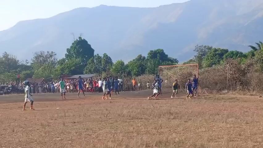 The video shows an outdoor soccer game being played on a dirt field. There are two teams of young men, one wearing green and white jerseys, the other wearing blue and white. A crowd of spectators watches from the sidelines, some standing and some sitting. In the background, there are trees and hills, and the sky is clear and bright. The game appears to be in progress, with players running and kicking the ball. One player in blue is attempting to kick the ball toward the goal, while the other team's players are trying to defend. The goal is a simple metal frame with a net. The overall scene suggests a community event, likely a local tournament or friendly match.