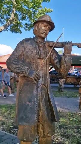 The video pans across a park-like setting featuring several wooden statues of musicians. The first statue is of a man playing a violin. The camera then moves to show a covered wooden structure with seating and more wooden carvings. Next, a statue of a man playing an accordion is visible. The video continues to pan, showing a statue of a man playing a drum. The final shot shows the same park setting with buildings in the background and people walking. The weather appears to be clear and sunny.