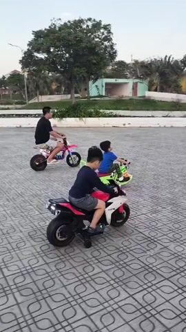 A man and two boys are riding on small, battery-powered vehicles in a park or paved plaza. The man rides a small motorcycle that is pink and green. One boy is in front on a red and white miniature motorcycle. The other boy is riding a small green toy car with a seat and backrest. All three are moving away from the camera. The area is paved with patterned tiles. In the background, there is a small bridge, trees, buildings, and several people walking. The sky is lightly overcast.