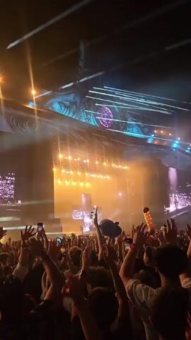 This is a vertical, first-person perspective video of a large crowd at an outdoor concert or festival. The crowd is dense and enthusiastic, with many people raising their hands in the air. Most individuals are facing towards a brightly lit stage in the distance. The stage features elaborate lighting rigs, large screens displaying abstract visuals, and prominent structures that resemble alien or futuristic architecture. Smoke and lasers are visible in the air, adding to the energetic atmosphere. The lighting is dynamic and changes color, primarily featuring warm yellows, oranges, and cooler blues and purples. The overall impression is one of excitement and immersion in a large-scale live entertainment event.