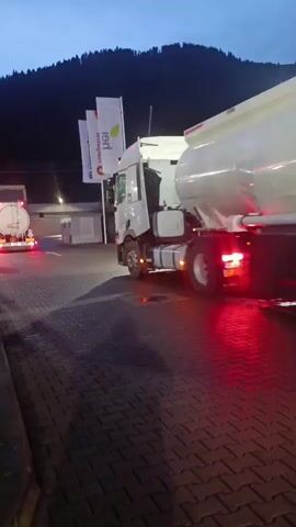A white tanker truck with a white cylindrical tank is parked on a paved area at dusk. The truck is facing away from the camera and is slightly angled to the left. Its rear lights are illuminated red. The truck has multiple axles and wheels. There are several tall, white flags with green logos visible to the left of the truck. In the background, a dark, tree-covered mountain is silhouetted against the twilight sky. Another white tanker truck is partially visible in the distance to the left, also parked. The ground is paved with interlocking bricks, and there appear to be some reflective surfaces or puddles on the ground.