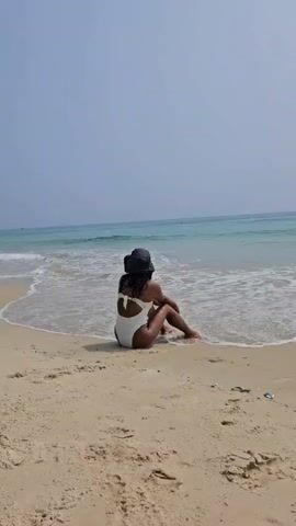 A video shows a person, likely a woman, from behind, sitting on a sandy beach at the edge of the water. She is wearing a white one-piece swimsuit with a cut-out back and a dark bucket hat. Her feet are partially submerged in the gentle waves that roll onto the shore. The ocean stretches out before her, displaying clear blue-green water under a bright, hazy sky. The sand is light brown with some footprints visible. A small piece of white and blue debris is visible on the sand to the right. The scene depicts a peaceful moment on a beach in Bac Cam Ranh, Vietnam, likely during the day.