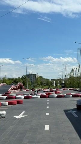 The video shows a go-kart track on a sunny day. The track is paved with grey interlocking tiles and bordered by red and white tires. There are white arrows painted on the track, indicating the direction of travel. Several go-karts are visible on the track, with drivers wearing helmets. In the background, there are trees, buildings, and a Ferris wheel. The sky is blue with scattered white clouds.