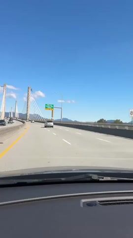 A forward-facing in-vehicle perspective shows a multilane highway bridge under clear blue skies. The Alex Fraser Bridge is visible, a cable-stayed structure with tall concrete pylons and many white suspension cables. Several cars are traveling on the bridge in both directions. A green highway sign indicates "113B Ave Airport Way 1700 m Exit Only." A tall industrial structure, possibly a smokestack, is visible on the right side of the bridge. The dashboard of the vehicle is visible at the bottom of the frame. The highway is marked with yellow lines on the left and white lines dividing lanes.