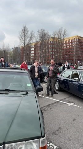 The video depicts a gathering of people in a parking lot, with classic cars on display. A man wearing a leather jacket is speaking into a microphone, seemingly addressing the crowd. He is standing near a dark green Volvo with the license plate "T015TT 178 RUS." Several other people are standing around, observing and listening. There's a blue Jaguar parked next to the Volvo. In the background, there is a large modern building with an orange and grey facade. The weather appears overcast.