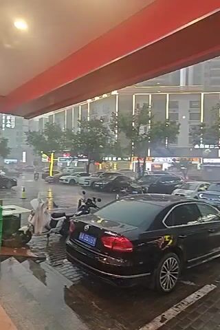 This is a first-person perspective video taken from an indoor overhang looking out onto a street during a heavy downpour. The street is wet and reflects the lights of nearby buildings and streetlights. Several cars are parked on the street, and some are driving through the rain. Two scooters are visible in the foreground, one parked and one potentially being ridden by a delivery person. The buildings in the background appear to be multi-story structures with illuminated windows and signs. Trees are also present along the street, their leaves appearing wet and possibly being blown by the wind. The overall impression is of a busy urban environment experiencing severe weather.
