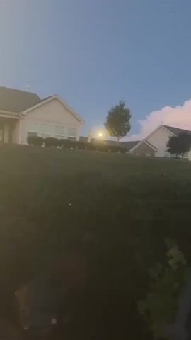 The video shows a row of houses on a grassy hill under a twilight sky. The sky is a deep blue with scattered clouds that are lit by the setting sun, casting pink and white hues. Several houses, appearing to be part of a residential development, are visible on the crest of the hill. They are uniformly designed, with light-colored siding and darker roofs. Each house has multiple windows and some have illuminated exterior lights. Trees are sparsely scattered on the property. The foreground shows a dark, out-of-focus area of vegetation and what appears to be a dark railing or fence. The camera is panning slowly to the right, revealing more of the houses and the landscape. There are no people or any discernible actions taking place in the video. The overall impression is one of a quiet, suburban evening.