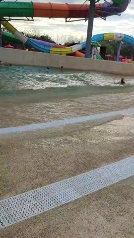 The video shows a wave pool at a water park. In the foreground, there is a sandy area with a white textured border. The wave pool itself is filled with water, and several people are visible swimming and playing in it. In the background, there are large, colorful water slides, indicative of a water park setting. The sky appears overcast. The waves in the pool are gentle, suggesting the wave-making mechanism is either off or operating at a low setting. People are casually enjoying the water, with some appearing to be children. A man in blue shorts stands in the water near the foreground, facing away from the camera. The overall scene depicts a leisurely day at a water park.