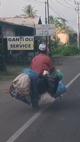 A person on a motorcycle is seen from behind, riding on a paved road. The person is wearing a red shirt and a white cap. The motorcycle is heavily loaded with various bags and bundles of goods, some of which appear to be textiles, plastic bags, and other items. The bags are piled high around the rider and secured to the rear of the motorcycle. The road appears to be in a semi-urban or rural area, with buildings and vegetation visible in the background. There is a sign on a building that reads "GANTI OLI DAN SERVICE," indicating a service station. The overall impression is of someone transporting a significant amount of goods on their motorcycle.