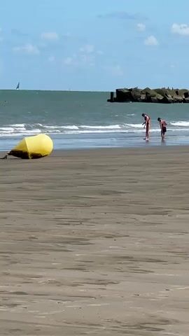 The video shows a wide shot of a beach with a yellow buoy in the foreground. Two people, likely children or teenagers, are standing in the shallow water near the shoreline, facing away from the camera and looking down. In the background, the ocean stretches out with gentle waves and a breakwater made of large rocks. A sailboat can be seen in the distance on the left side of the frame. The sky is blue with a few scattered clouds. The sand on the beach is wet in the foreground and drier further back. The overall scene depicts a peaceful day at the beach with two individuals exploring the water's edge.