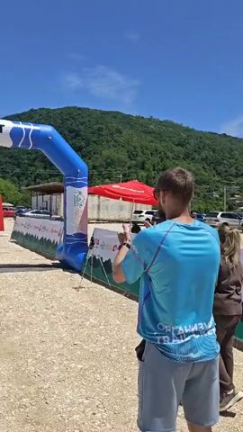 A man in a green t-shirt, black shorts, and a white cap runs under a blue and white inflatable arch marked "START." He stops and checks his watch. Another man in a light blue t-shirt stands to his right, holding a phone. A third man in a black t-shirt approaches the runner, holding a microphone. The scene is outdoors on a dirt path, with green mountains in the background under a clear blue sky. White and red flags line the path, and several vehicles and tents are visible further back.