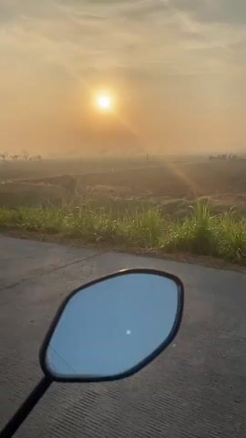 The video shows a sunrise over a rural landscape. The sun is low in the sky and appears hazy, casting a warm glow over the scene. The foreground features the side mirror of a motorcycle, reflecting the sky and the surrounding environment. Beyond the mirror, a road can be seen, with green vegetation along the sides. In the distance, fields are visible, some appearing to be cultivated land, with a few scattered trees and structures. There are also a few people and vehicles, including a bicycle and motorcycles, in the distance. The overall atmosphere is peaceful and serene, conveying the tranquility of a rural morning.