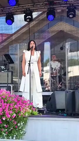 A woman in a white jumpsuit stands on a stage, singing into a microphone. She has long, dark hair and is wearing jewelry. Behind her, a man plays drums. In front of her are pink flowers and speakers. The stage is set up outdoors, with a large screen or backdrop behind the performers. The lighting is blue and bright.