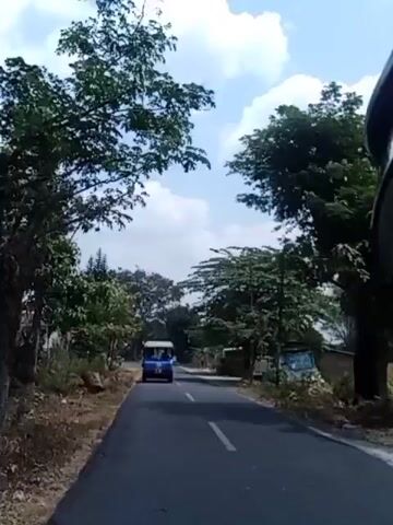 A blue vehicle is driving down a paved road, surrounded by trees and vegetation. The sky is visible overhead with clouds. The vehicle appears to be a small van or bus. The road has a white line marking the center.