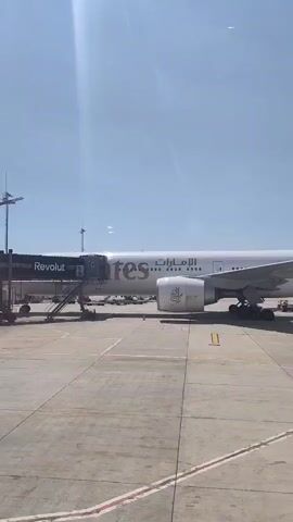 A commercial airplane is parked at an airport gate. A jet bridge is connected to the airplane. Another airplane is in the sky in the background. The sky is clear and blue. The scene is captured from inside a building, possibly a terminal, looking out a window. The text on the airplane reads "Emirates". The text on the jet bridge reads "Revolut".