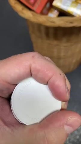 A close-up shot focuses on a hand holding a miniature jar of Nutella. The hand, with visible skin texture and a ring on the ring finger, grips the small glass jar. The jar is topped with a white plastic lid and has a label clearly displaying the "nutella" logo in red and white. Below the logo, the text "25 g e" is visible. A barcode is also present on the label. The jar appears to be in a wicker basket filled with various chocolate bars and other food items. In the background, blurred shelves of a supermarket can be seen, with colorful products lining them. The lighting suggests it is daytime, likely indoors in a well-lit store. The overall tone is casual and exploratory, possibly someone browsing or selecting items in a grocery store in Friedrichshafen, Germany.
