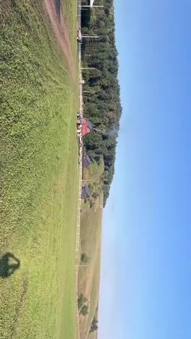 The video shows a rural landscape with a large grassy field in the foreground. On the right side, there is a wooded hillside with a clear blue sky above. In the middle ground, there are several small, modern cabins with dark roofs and solar panels. A white fence runs along the edge of the field, separating it from the cabins. At the top of the frame, near the wooded area, there are some buildings, including a red-roofed structure that appears to be a house or barn. A blue tractor is parked near this building, and there are a few other vehicles, possibly ATVs, lined up behind it. Smoke is rising from the chimney of the red-roofed building. The video is filmed from a high vantage point, possibly from a drone or a hot air balloon, as a shadow in the shape of a balloon gondola is visible in the lower left corner of the frame. The overall scene suggests a quiet, rural setting, possibly a farm or a holiday resort.