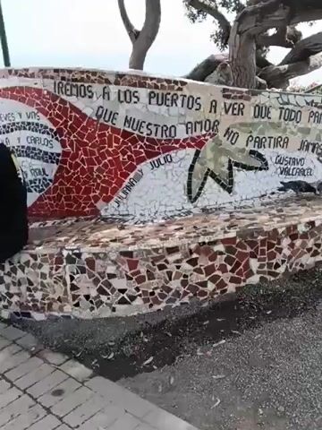 The video shows an outdoor scene with a long, winding mosaic bench. The bench has various designs and lettering on it, including names and quotes in Spanish. Several people are present. Some individuals are sitting on the bench, while others are standing around it, taking photos with their phones. A large, gnarled tree provides shade near the bench. The background shows a seaside landscape with buildings in the distance. The overall atmosphere appears relaxed and touristy, with people enjoying the scenery and taking pictures. The ground is covered with small pebbles and debris.