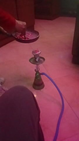 In a close-up shot, a person in a red shirt is using tongs to place burning charcoal from a metal pan onto the bowl of a hookah, which is sitting on a tile floor. The hookah is green and silver, with a blue hose attached. The room is lit with a pink light. A pair of white sneakers and a person's leg clad in dark pants are visible in the periphery.