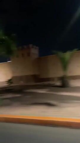 The video shows a night drive through an urban area, likely Mechouar Kasbah, Morocco, as viewed from inside a moving vehicle. The scene is illuminated by streetlights, creating significant lens flares throughout the footage.

The video begins showing buildings with traditional Moroccan architecture, characterized by light brown or tan walls and distinct window shapes, along with palm trees. A long, fortified-looking wall is visible in the initial frames. As the vehicle moves, the interior of the car (dashboard and side mirror) can be seen in the bottom left corner.

A building with the text "JANA" clearly visible on its facade comes into view, featuring multiple small, arched windows. The surrounding area includes paved walkways and low walls, suggesting a public square or street. Further along, a section of the street and some vegetation are bathed in a vibrant purple light, creating a striking contrast with the otherwise warm-toned lighting. The motion blur from the moving vehicle is consistent throughout the video.