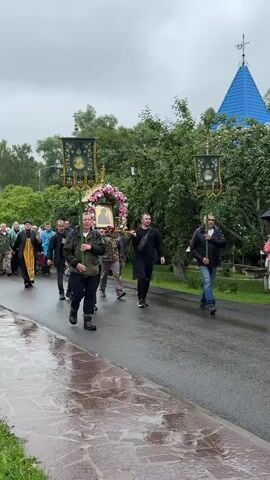 A group of people, including a priest, are walking in a procession on a wet road. The priest is wearing a black and yellow robe. Several people are carrying banners and a decorated structure with an icon. The procession is moving from left to right in the frame. In the background, there are trees and a blue-domed building. The sky appears overcast.