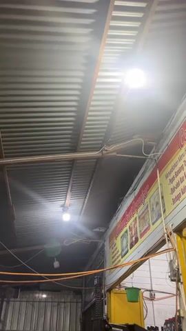 The video pans across a dimly lit workshop. The ceiling is corrugated metal, supported by exposed beams and girders, with several fluorescent lights illuminating the space. Wires and orange hoses are strung across the ceiling. On the right, a colorful sign with text and images is mounted on the wall. Below the sign, a yellow structure, possibly a machine, is visible, along with shelves holding various bottles and containers. In the center of the frame, a large, dark piece of equipment dominates the foreground, with a blue base and various metal components. To the left, a motorcycle is parked, with red chairs scattered around the area. The floor appears to be concrete and is somewhat cluttered. The overall impression is of a functional, albeit somewhat unkempt, automotive repair shop.