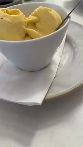 The video opens with a close-up of a white bowl containing three scoops of yellow ice cream, served on a white plate with a white napkin. A metal spoon is inserted into the ice cream. In the background, a white table is visible with several items, including two salt and pepper shakers, a glass bottle of water with a blue label, a green glass bottle with a yellow label, an ashtray, and part of another plate with cutlery. The scene appears to be indoors, possibly a cafe or restaurant, with a hint of outdoor greenery in the upper left corner. The camera then pans slightly to the right, revealing a woman with curly brown hair and a white lace top sitting at the table. She is looking down and to the side, seemingly engaged in conversation or activity. The final seconds of the video show her looking more directly towards the camera with a slight smile. The audio contains a voice stating "I can't wait to try this ice cream of cactus" and "Cactus ice cream."