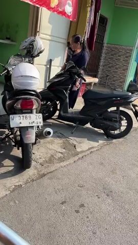 A woman stands next to a black motorcycle. She is wearing a blue shirt and has her hair pulled back in a ponytail. She is holding a phone to her ear and appears to be speaking. Two motorcycles are parked side-by-side. The motorcycle on the right is black. The motorcycle on the left is white and gray. Both motorcycles have helmets on them. The scene appears to be outdoors on a street. There is a building in the background.