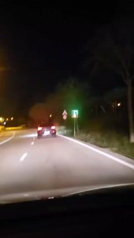 The video shows a car driving on a dark road at night. The car is following another vehicle, which is visible ahead. There are streetlights along the road, and trees line the side of the road. A digital speed limit sign displays 33, next to a traditional speed limit sign of 60. The road has lane markings. The view is from the driver's perspective, looking through the windshield. The overall lighting is dim, with the car's headlights illuminating the road.