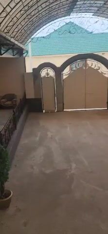 The video shows a courtyard with a covered walkway leading to a gate. The courtyard is paved with concrete, and there is a small potted plant in the lower left corner. To the left, there is a sofa under the covered walkway. The gate is large and ornate, with a smaller door on either side. The gate is closed, and it is unclear if there is anyone behind it. The background shows a building with a green tiled roof. The video is shot from a slightly elevated perspective, looking down into the courtyard.