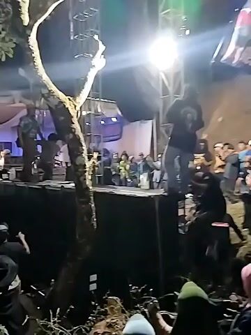 A group of people are gathered at night in an outdoor space. A band is performing on a raised stage. The audience is standing and watching the band, some are dancing. There are trees and lights in the background. The scene appears to be a concert or festival.