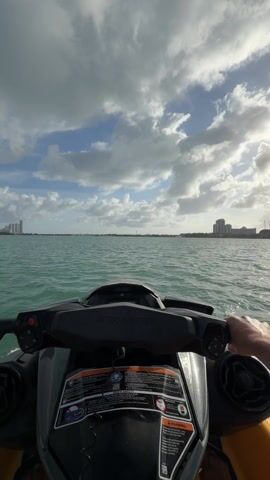 A person is riding a Sea-Doo jet ski on a body of water. The camera is mounted on the jet ski, providing a first-person perspective. The sky is partly cloudy with sunlight breaking through. In the distance, a cityscape is visible. The water is turquoise, and there are small waves. The rider's hands are visible on the handlebars.