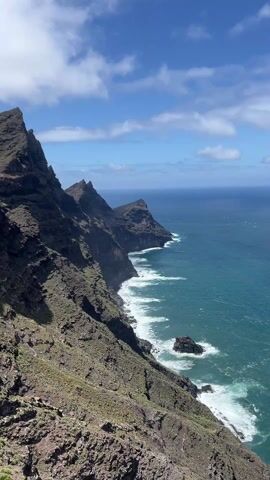 The video shows a mountainous, rocky coastline and then a group of people on a glass observation deck. The rocky coastline features steep cliffs, and white waves crash against the rocks below. The sea is a deep blue-green. The sky is blue with scattered white clouds. A group of about seven people stands on a glass-floored observation deck, looking out at the view. Some are taking pictures or looking at their phones. The deck appears to be built into the side of the mountain.