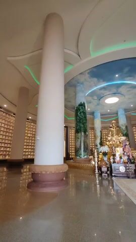 The video shows the interior of a Buddhist temple.  The floor is shiny and reflective.  Tall white pillars support the ceiling, which features a circular design painted with a blue sky and clouds, accented by blue and green lighting.  A golden Buddha statue sits at the center, surrounded by offerings on a decorated table.  The walls are lined with what appear to be individual niches, possibly for remains or memorial items. No people are visible.
