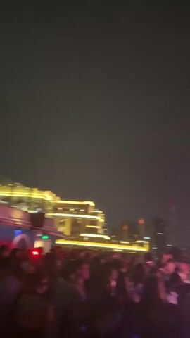 The video shows a night scene in a city with a crowd of people gathered. In the background, there are illuminated buildings and a tower. The scene is lit by colorful lights, predominantly pink and red, which create a party or event atmosphere. There are some visual effects like light beams. A flag on a flagpole is also visible. The camera is panning to show the cityscape and the gathering.