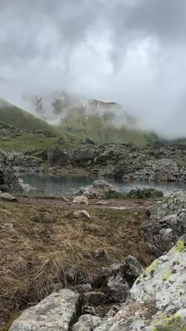 This media file depicts a serene natural landscape in Blo, Georgia. The scene features a tranquil lake nestled amidst rugged, rocky terrain. In the background, verdant, rolling hills ascend towards mist-shrouded mountains, their peaks partially obscured by clouds. The weather appears overcast and possibly damp, with a soft, diffused light characteristic of a cloudy day. The foreground is dominated by dry, golden grasses and large, moss-covered rocks, suggesting a wild and untamed environment. The overall mood is one of peaceful solitude and the raw beauty of nature. There are no people or objects of human creation visible in the frame. The natural elements – the lake, rocks, grass, and mountains – are the sole subjects. The time of day is indeterminate due to the overcast sky, but the lighting suggests it could be any time from late morning to late afternoon.