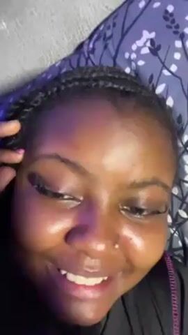 A close-up shot shows a woman with dark skin, braided hair, and a nose piercing lying on a patterned pillow. Her eyes are closed, and she is seen winking, and rubbing her eyes with a fingernail painted pink. She smiles slightly at the beginning, and a tear falls during the video. She is wearing what looks like a black shirt. The lighting in the room appears to be from a purple light source.
