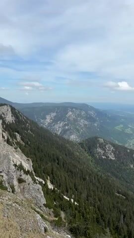 The video presents a panoramic view from a mountainous area. The foreground features rocky terrain and evergreen trees sloping down into a valley, with a winding path visible on the left side. In the distance, more mountain ranges stretch out into the horizon, and a small town is nestled in the valley below. The sky is partly cloudy with blue patches visible.  The camera pans slowly to the right, providing a broader view of the landscape.