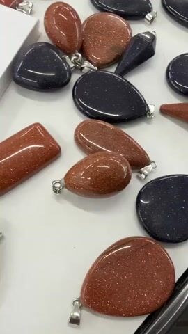 On a white surface are several pendants of different shapes and colors with metal loops. The pendants are made of glass. There are teardrop, oval, heart, and rectangular shapes. Some are reddish-brown, and some are dark blue. The pendants have a sparkling, glittery appearance. In the upper right corner, there is a white cardboard box with text in Russian that says "Aventurine glass". The box also indicates the price of 1 piece is 1-500 rubles. The camera pans slowly over the collection of pendants.