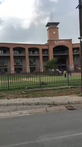 The video shows a three-story brick building with a clock tower on top, labeled "SPARK." A black metal fence separates the building and a grassy area from the street. A few people can be seen walking on the grass. Cars pass on the street in front of the building. The sky is overcast.