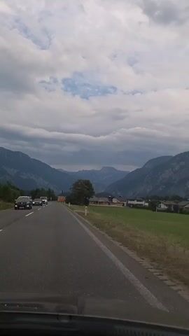 The video captures a point-of-view shot from inside a vehicle traveling on a paved road. The road is lined with white and yellow markers. On the left side of the road, there is a grassy area with trees and a few cars can be seen driving in the same direction. On the right side of the road, there is a wide grassy field, and in the distance, there are houses and buildings. The background is dominated by large mountains under a cloudy sky. The weather appears overcast.