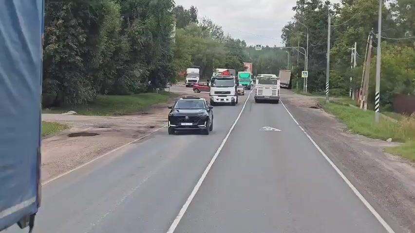 The video shows a view from a vehicle traveling on a two-lane road. The road is wet, indicating recent rain. A black SUV is in the foreground, in the right lane. Several trucks, including a white Volvo with a crane, a green panel van, and a white bus, are traveling in the same direction in both lanes. A red car is stopped in the left lane. The surroundings are green and wooded, with some buildings visible on the left. There are pedestrian crossings and traffic lights ahead. The sky is overcast.