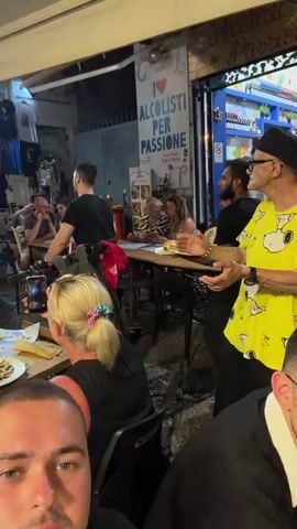 This video captures an outdoor dining scene at night, possibly a street market or restaurant in a European location, given the signs in Italian. The foreground shows several people seated at tables, with plates of food and drinks, including wine and soda cans, on the tables. One woman in a red shirt is looking to her left, while another woman with blonde hair tied back with a colorful scrunchie is holding up her phone, seemingly recording or taking a photo. In the background, a man in a bright yellow t-shirt with a Snoopy graphic and a black hat, wearing glasses, is gesturing with his hands while speaking to someone off-camera. He appears to be the focus of attention for some patrons. Behind him, a sign reads "I <3 ALCOLISTI PER PASSIONE" which translates to "I love alcoholics with passion." Other individuals are visible at tables and walking around, some wearing black t-shirts. The environment is lit by string lights overhead and the illuminated storefronts behind the seating area, which display clothing and other merchandise. The overall atmosphere seems lively and social, with people enjoying a meal and entertainment.