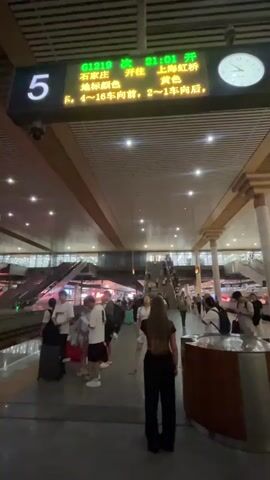 A video taken at a train station platform shows a digital display board hanging from the ceiling. The board displays train information, including departure times, destinations, and car numbers. Below the board, passengers are seen standing on the platform, some with luggage, waiting for their trains. Escalators and other station infrastructure are visible in the background. The overall scene depicts a busy train station environment.