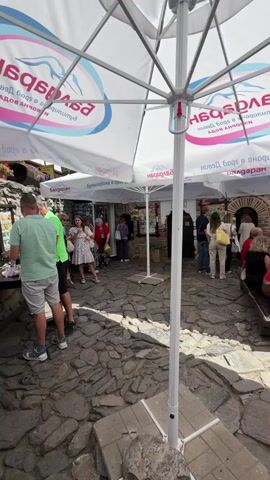The video shows an outdoor seating area with cobblestone paving. Several large white umbrellas with branding are set up, providing shade. In the foreground, two men are standing near a table, with one man in a green t-shirt and grey shorts looking down at something on the table. Behind them, a group of people, including women and children, are gathered. To the right, another group of women are seated at a wooden picnic table under one of the umbrellas, engaged in conversation. In the background, there are stone buildings and some greenery, suggesting a rustic or historical setting. A car is visible in the distance. The overall atmosphere appears to be a casual gathering in a public outdoor space.