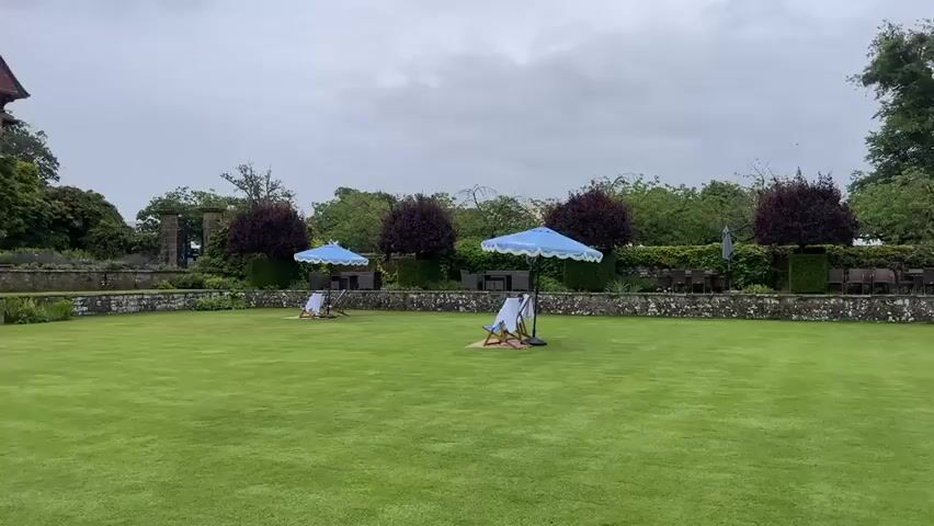 The video shows a large, well-manicured green lawn with several empty lounge chairs and blue and white umbrellas set up. In the background, there is a stone wall bordering the lawn, followed by a garden area with manicured bushes and trees. Beyond the garden, a large, historic stone building with a red tile roof and multiple windows can be seen. The sky is overcast with clouds. The camera pans slowly from left to right, offering a panoramic view of the grounds and the building.